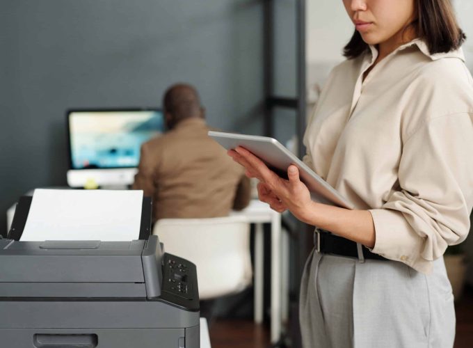 Focus on tablet held by young elegant businesswoman computing in office while standing by xerox machine against African American male coworker